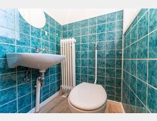 Kleines Badezimmer mit türkisfarbenen Wandfliesen, wandmontiertem Waschbecken und WC. An der Rückwand steht ein weißer Heizkörper; der Boden ist in Holzoptik gehalten.