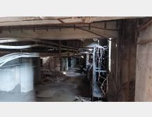 Ein verlassenes Kellergeschoss mit sichtbarer Stahlbetonkonstruktion, herabhängenden Kabeln und Rohren sowie herumliegendem Schutt auf dem Boden. Die Umgebung wirkt dunkel und ungenutzt, mit teilweise beschädigten Bauelementen.