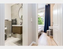 Blick durch einen Flur in ein modernes Badezimmer mit grauen Steinfliesen, wandhängendem Waschtisch und rundem Spiegel sowie in ein angrenzendes Schlafzimmer mit großem Fenster, dunklen Vorhängen und hellem Holzboden. Neben dem Bett stehen ein kleiner Schreibtisch und ein gepolsterter Hocker.