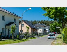 Ein ruhiges Wohngebiet mit freistehenden Einfamilienhäusern, Vorgärten und Zäunen entlang der Straße. Im Vordergrund ein parkendes Auto und ein Straßenschild mit der Aufschrift „Bachallee“, im Hintergrund ein Waldgebiet.