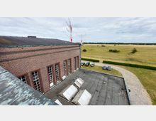 Backsteingebäude mit großen Sprossenfenstern und Flachdach mit Lichtkuppeln, umgeben von einer gepflasterten Einfahrt und parkenden Autos. Im Hintergrund eine weitläufige Wiesenlandschaft und rote Antennenmasten vor einem bewölkten Himmel.