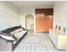 Wohnzimmer mit hellem Holzboden, einer großen braunen Eckcouch mit Kissen und einem Teppich in der Mitte. Im Hintergrund sind ein weißer Durchgangsbogen, ein geschlossener Holzschrank und eine helle Kommode mit dekorativen Bildern zu sehen.