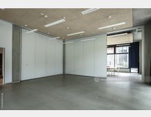 Ein moderner, leerer Innenraum mit glatten Betonböden, weißen Trennwänden und einer abgehängten Decke mit integrierter Beleuchtung. Im Hintergrund befindet sich ein Bereich mit großen Glasfenstern, die auf einen Außenbereich blicken, sowie ein Tisch mit Stühlen.