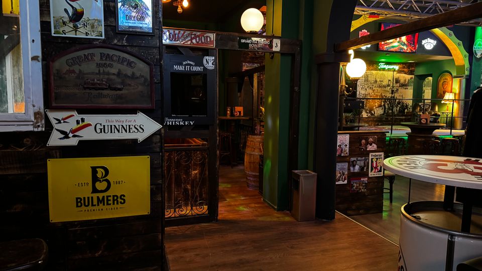 Innenraum einer irischen Pub-Bar mit dunkler Holzvertäfelung, vielen Vintage-Werbeschildern (u. a. Guinness und Bulmers) und warmen Kugellampen. Ein Durchgang führt zu einem Sitzbereich mit hohen Tischen, grün gestrichenen Bögen und beleuchteten Bar-Schildern an den Wänden.