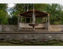 Steinerner Brunnen mit einer Statue in der Mitte, umgeben von einem halbkreisförmigen Pavillon mit Säulen und einem verzierten Holzdach, eingebettet in eine Gartenanlage mit symmetrischen Blumenbeeten. Im Hintergrund dichter Baumbestand.