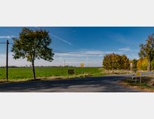 Landstraße mit einer Kreuzung, flankiert von Bäumen und Stromleitungen, im Hintergrund eine grüne Wiese mit mehreren Windkraftanlagen unter einem blauen Himmel. Straßenschilder und ein Ortseingangsschild weisen auf nahegelegene Ortschaften hin.