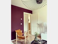 Ein modernes Wohnzimmer mit einer violetten Akzentwand, einem Holzsessel mit Ledergurten und einem runden Couchtisch mit Glasplatte. Im Raum stehen eine weiße Tür, eine transparente Tischlampe und eine Vase mit weißen Blumen.