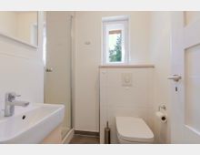 Kleines, helles Badezimmer mit weiß gefliesten Wänden, Waschbecken im Vordergrund, einer Glasduschkabine und einer wandhängenden Toilette. Über dem Spülkasten befindet sich ein schmales Fenster; der Boden ist dunkel gefliest.