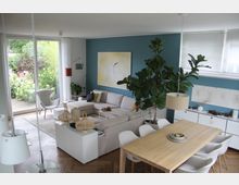 Modernes Wohnzimmer mit offenem Essbereich. Ein großes Ecksofa steht vor einer blauen Wand mit abstraktem Kunstwerk, ein Esstisch aus Holz mit weißen Stühlen befindet sich im Vordergrund, und raumhohe Fenster bieten Blick auf einen grünen Garten.