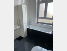 Modernes Badezimmer mit weißen Wänden und großen, dunkelgrauen Fliesen auf dem Boden. Der Raum enthält eine Badewanne, ein Waschbecken und einen hohen weißen Schrank; ein großes Fenster mit satiniertem Glas lässt Licht herein.
