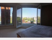 Blick aus einem Schlafzimmer mit einem Bett im Vordergrund durch weit geöffnete Glastüren auf eine Terrasse mit Topfpflanzen und einer Aussicht auf eine Stadtlandschaft, inklusive eines markanten Fernsehturms im Hintergrund. Links ist eine Backsteinmauer zu sehen.