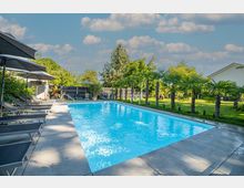 Außenbereich mit einem rechteckigen Swimmingpool, von Liegestühlen und Sonnenschirmen umgeben, mit Rasenflächen und kleinen Palmen im Hintergrund. Im Umfeld befinden sich Bäume und ein Wohnhaus, unter einem sonnigen, leicht bewölkten Himmel.