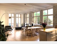 Loftartige Wohnung mit offenem Wohn- und Essbereich, weißen Sichtziegelwänden und großen Fenstern, die viel Tageslicht hereinlassen. Der Raum ist mit Holzböden, modernen Möbeln, einem Esstisch mit Holzstühlen und einer Kücheninsel ausgestattet.