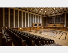 Innenansicht eines Konzertsaals mit gestuften Sitzreihen, Holzvertäfelung, einer gewölbten Bühne und einer großen Orgel auf der Rückwand. Die Wände sind mit hohen Säulen und Gittern verziert, während die Decke mit quadratischen Paneelen gestaltet ist.