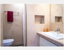 Beigefarbenes Badezimmer mit Wandfliesen, einem weißen Waschbecken mit silbernem Wasserhahn und zwei orangefarbenen Bechern darauf. Links befindet sich eine Toilette, ein Handtuchhalter mit einem roten Handtuch und eine Duschvorhangstange.