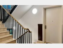 Treppenhaus eines modernen Wohngebäudes mit einer weißen Wand, schwarzen Geländern und beigen Marmorstufen. Rechts befindet sich eine weiße Wohnungstür mit Türspion und Metallschloss, darüber eine runde Wandlampe.