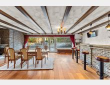 Gemütlicher Aufenthaltsraum mit Holzboden, sichtbaren Deckenbalken und Backsteinwänden, ausgestattet mit Esstisch und Stühlen sowie einer langen Bar mit Barhockern. Große Glas-Schiebetüren mit roten Vorhängen öffnen den Blick in einen Gartenbereich.