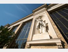 Fassade eines historischen Gebäudes mit großen Fenstern und gelbem Backstein. An der Wand befindet sich eine weiße Skulptur in einer Nische, die eine stehende Figur darstellt, umgeben von blauen Himmel und grünem Laub am unteren Bildrand.