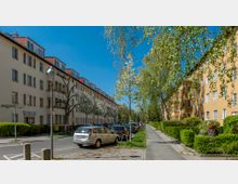 Wohnstraße mit mehrstöckigen, gelb und cremefarbenen Wohngebäuden mit roten Ziegeldächern, flankiert von Bäumen und gepflegten Hecken. Am Straßenrand sind geparkte Autos, und ein Gehweg führt entlang der Grünanlagen.