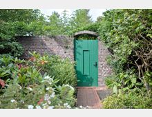 Ziegelmauer mit einer kleinen, grünen Holztür, umgeben von üppigem Garten mit verschiedenen Büschen, Blumen und dichtem Laub; ein schmaler Ziegelweg führt zur Tür.