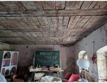 Ein rustikaler Innenraum mit abblätternden Wänden und einer grob verarbeiteten Holzdecke. Im Raum befinden sich eine Tafel mit der Aufschrift „Markt Treff ab August 25“, ein Regal, Möbelstücke und dekorative Gegenstände, sowie kleine Fenster mit Rundbögen.