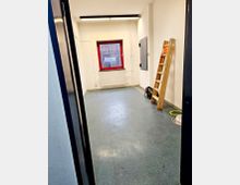 Ein schmaler Raum mit weißen Wänden und einem grauen Bodenbelag. Auf der rechten Seite steht eine Holzleiter, und vor einem roten Fenster mit Blick nach draußen befindet sich ein weißer Heizkörper. Kabel und eine Metallklappe an der Wand sind ebenfalls sichtbar.