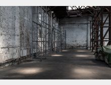 Innenansicht einer leerstehenden Industriehalle mit weißen, teils verwitterten Backsteinwänden, einer abgenutzten Betonbodenfläche und Stahlgerüsten an den Seiten. Die Halle hat ein offenes Holztragwerk an der Decke, und im Vordergrund liegt eine plane grüne Abdeckung über gelagerten Objekten.