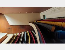 Innenraum eines Auditoriums mit bunten Sitzreihen, die von unten nach oben in Halbrundform angeordnet sind. Die Wände sind teilweise mit Holzvertäfelung und glatten, hellen Flächen gestaltet; an der Seite führt eine Treppe hinauf.