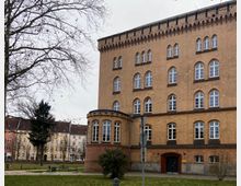 Backsteinbau mit mehreren Stockwerken und hohen, symmetrischen Fenstern sowie einem runden Erker an der Seite; im Vordergrund eine Grünfläche mit Bäumen, Parkbänken und Gehwegen, im Hintergrund mehrstöckige Wohnhäuser.