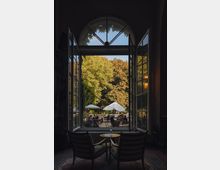 Blick durch ein großes, geöffnetes Bogenfenster auf eine Terrasse mit Sitzplätzen und weißen Sonnenschirmen, umgeben von Bäumen. Im Vordergrund befinden sich zwei Sessel und ein kleiner runder Tisch in einem Innenraum.