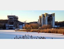 Industrielle Wasserstraße mit einem Schiffhebewerk im Vordergrund, bestehend aus einer Stahlkonstruktion und modernen Gebäuden mit gelben Akzenten; umgeben von schneebedeckten Landschaften, kahlen Bäumen und vereistem Wasser.