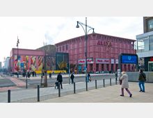 Einkaufszentrum "Alexa" mit pinkfarbener Fassade und markantem Schriftzug, umliegende Geschäfte wie MediaMarkt und H&M; im Vordergrund Straßenkreuzung mit Personen sowie ein Eingang zur U-Bahn-Station "Alexanderplatz".