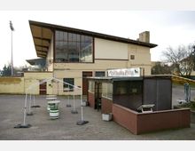 Ein flacher gelber Backsteinbau mit großen Fensterfronten auf der oberen Etage, daneben ein überdachter kleiner Imbissstand mit der Aufschrift „Zur Traber-Pause“. Im Vordergrund stehen einige Stehtische und weiße Metallgestelle auf einem Asphaltplatz.