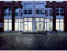 Moderne Gebäudefassade mit großen Fensterelementen und einer Eingangstür, auf der "ARZT" steht. Im Inneren des Gebäudes sind beleuchtete, helle Räume zu sehen, die eine Arztpraxis oder ähnliche Einrichtung nahelegen.