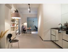 Moderner Büroraum mit minimalistischer Einrichtung. Links ein Regal mit Büchern und Dekorationen, rechts eine kleine Küchenzeile mit einer Kaffeemaschine auf der schwarzen Arbeitsplatte, im Hintergrund Schreibtischarbeitsplätze mit Stühlen und Lampen.