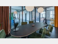 Ein moderner Besprechungsraum mit einem ovalen Tisch, grünen Stühlen und einem Computerbildschirm darauf. Im Hintergrund sind durch Glaswände angrenzende Räume sichtbar, mit hellen Wänden, großen Fenstern und dekorativen Elementen wie Vorhängen und Pflanzen.