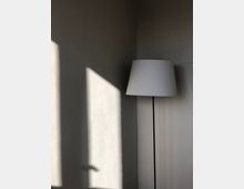 Eine schlichte Stehlampe mit weißem Lampenschirm steht in einer Ecke vor einer hellen, beige gestrichenen Wand. Sonnenlicht fällt durch ein Fenster und wirft geometrische Schatten auf die Wand.
