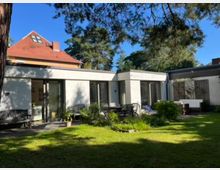 Einfamilienhaus mit einem flachen, modernen Anbau und einer vorgelagerten Terrasse, umgeben von einem Garten mit Rasen, Pflanzen und Bäumen; im Hintergrund ein traditionelles Haus mit rotem Satteldach.