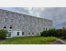 Ein mehrstöckiges Gebäude mit grauer, strukturierter Fassade und zahlreichen rechteckigen Fenstern. Im Vordergrund befinden sich Grasflächen, Büsche und ein großer Stein, während der Himmel bewölkt ist.