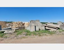 Ein offenes Lagergelände mit gestapelten Betonblöcken und großen Haufen aus Holzabfall, umgeben von Grünflächen und einem klaren blauen Himmel. Im Hintergrund sind Bäume und einige Stromleitungen sichtbar.