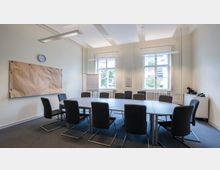 Ein schlicht eingerichteter Konferenzraum mit einem ovalen Tisch und elf gepolsterten Stühlen. Im Raum befinden sich zwei Fenster, eine Pinnwand, ein Flipchart, eine Uhr an der Wand und ein kleiner Schrank mit Kaffeemaschine.