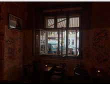 Innenraum eines kleinen Cafés mit dicht beschmierten, graffitiartigen Wänden und dunklen Holztischen mit Stühlen. Ein großes, teilweise geöffnetes Fenster mit Oberlichtern zeigt den Blick auf die Straße mit geparkten Autos und gegenüberliegenden Gebäuden.