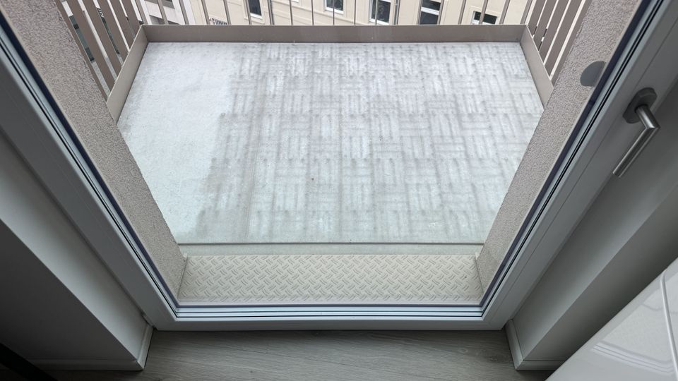 Blick durch eine Glastür auf einen kleinen Balkon mit hellgrauem Betonboden und umlaufendem Metallgeländer. Im Hintergrund sind die Fassaden und Fenster eines benachbarten Gebäudes zu sehen.