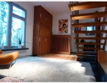 Wohnraum mit modernem Design: Links ein großes Fenster mit Blick ins Freie, daneben ein Holzschrank, eine Heizung mit Holzverkleidung und ein Gemälde. Rechts schwebende Holztreppenstufen, ein heller Teppich liegt auf dem dunklen Boden.