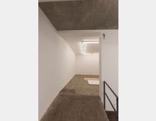 Minimalistischer Innenraum mit weiß gestrichenen Wänden, Betonboden und einer Betondecke. Im Hintergrund ist ein rechteckiges, integriertes Deckenlicht und ein kleiner Bereich mit Teppichboden sichtbar.