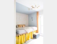 Schlafzimmer mit einem erhöhten Bett auf hölzernen Beinen, bedeckt von heller Bettwäsche und gelben Vorhängen, die den Raum darunter verdecken. Im Hintergrund eine hellblaue und weiße Wand, gelbe Vorhänge vor einem großen Fenster, und eine moderne Deckenleuchte.