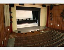 Moderner Veranstaltungsraum mit Blick auf eine Bühne mit schwarzen Vorhängen und hellem Hintergrund; davor befinden sich ansteigende Sitzreihen und hölzerne Wandverkleidungen mit eingebauten Lautsprechern und Beleuchtung.