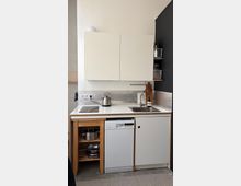 Kompakte Küchenzeile mit weißen Hängeschränken, Kochfeld und Edelstahlspüle auf einer weißen Arbeitsplatte sowie einem Geschirrspüler darunter. Rechts sind offene Regale an einer dunkelgrauen Wand und ein metallischer Spritzschutz zu sehen.