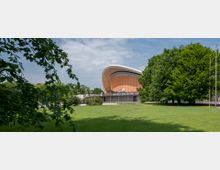 Moderne Architektur eines Gebäudes mit geschwungenem Dach und einer Fassade aus orangefarbenen Paneelen, umgeben von einer weitläufigen Rasenfläche und Bäumen im Vordergrund. Ein Schriftzug am Gebäude verweist auf eine Ausstellung, und der Himmel ist leicht bewölkt.