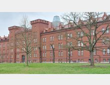 Backsteinbau mit historischer Architektur und zahlreichen weißen Rundbogenfenstern, umgeben von einer Grünfläche mit einigen kahlen Bäumen im Vordergrund. Ein gläserner Aufbau befindet sich im Dachbereich des Gebäudes.